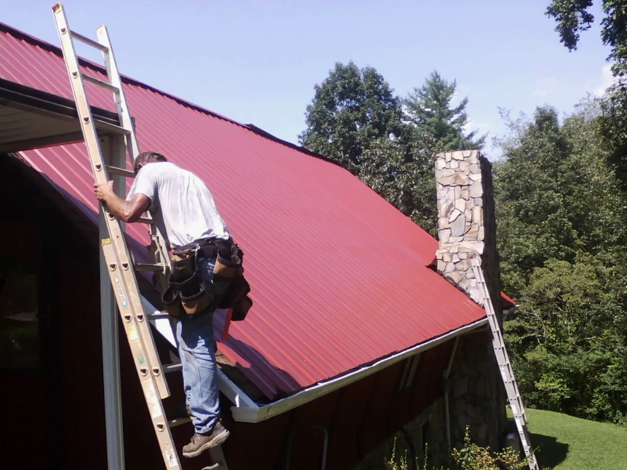Roofer on a ladder beside a red metal roof and stone chimney — Soffit & Fascia Repair by Roofing Brothers in Meyersdale